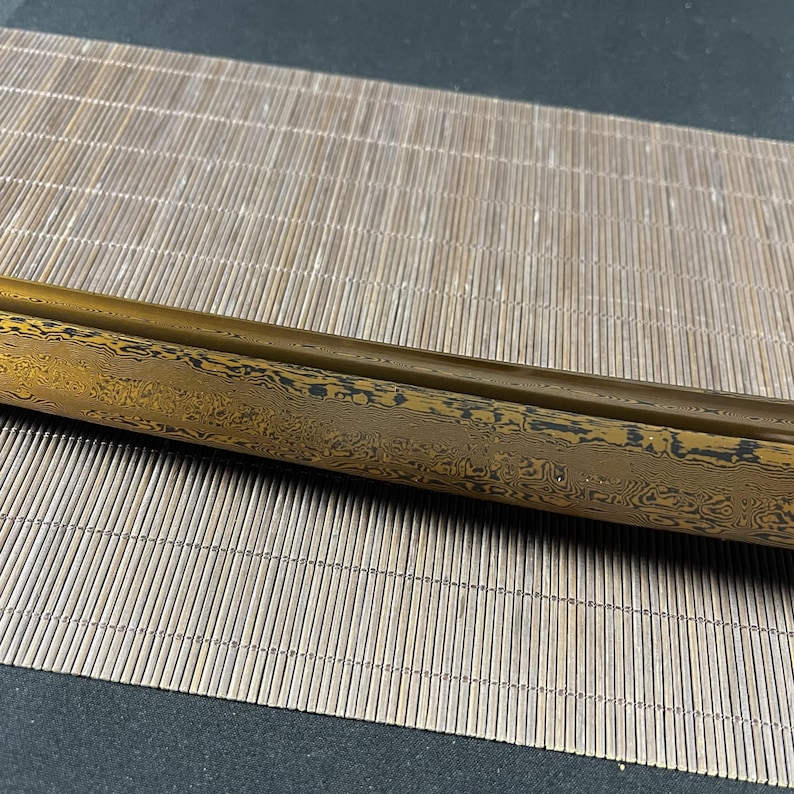 May include: A close-up of a gold and black sword blade with a wavy pattern. The blade is resting on a woven bamboo mat.