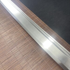 May include: Close-up of a silver sword blade with a polished finish. The blade is resting on a brown bamboo mat.