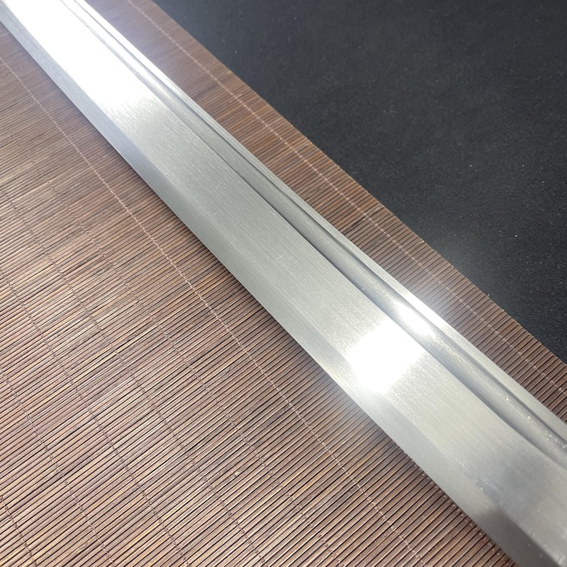May include: Close-up of a silver sword blade with a polished finish. The blade is resting on a brown bamboo mat.