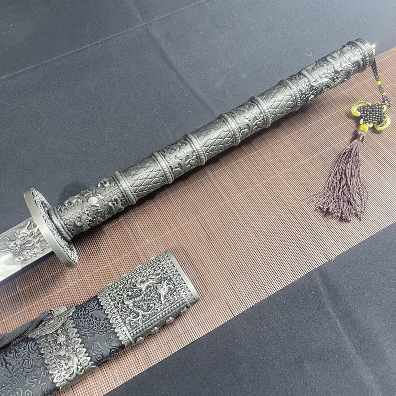 May include: A silver-coloured sword with intricate carvings and a black scabbard. The sword has a tassel attached to the handle.