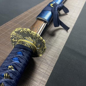 May include: A close-up of a katana sword with a blue and gold scabbard. The scabbard is decorated with a wave pattern and the kanji for "sword".