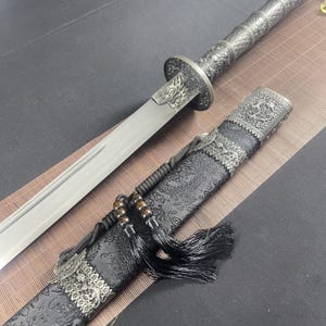 Puede incluir: Una espada plateada y negra con un diseño de dragón en el mango y la vaina. La vaina está cubierta de una tela negra con un patrón de dragón. La espada tiene un borla negra.