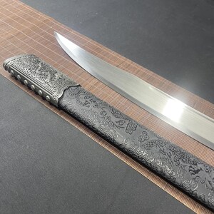 May include: A silver sword with a black scabbard featuring an intricate dragon design. The scabbard has a silver metal tip and a silver metal band with decorative accents.