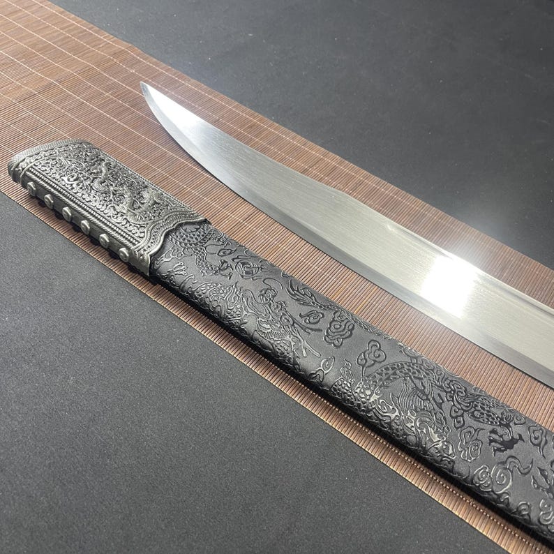 May include: A silver sword with a black scabbard featuring an intricate dragon design. The scabbard has a silver metal tip and a silver metal band with decorative accents.