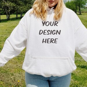 May include: White hoodie with a front pocket and the text "YOUR DESIGN HERE" in black. The hoodie is made of a soft material and is suitable for custom designs. The model is wearing blue jeans.