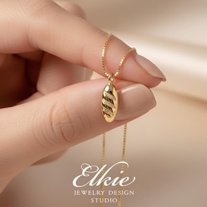 Peut inclure: Un collier doré avec un petit pendentif ovale présentant une surface texturée à rayures diagonales. Le pendentif est suspendu à une fine chaîne de type boîte. Le texte 'Elkie JEWELRY DESIGN STUDIO' est imprimé en blanc au bas de l'image.