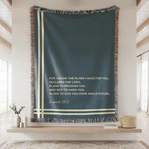 May include: A woven teal throw blanket with cream-colored stripes and fringe. The blanket features the text "FOR I KNOW THE PLANS I HAVE FOR YOU...Jeremiah 29:11" in white. The blanket is displayed on a wooden shelf.