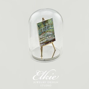 May include: A miniature painting of a bridge over a pond with water lilies, displayed on a small easel under a glass dome.