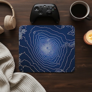 May include: A navy blue mousepad with a white topographical map design. The mousepad is surrounded by a black game controller, a cup of coffee, a lit candle, and a pair of headphones. The items are on a dark wooden surface.