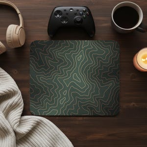 May include: A dark green mousepad with a topographic map design in light brown. The mousepad is surrounded by a black game controller, a cup of coffee, a lit candle, and headphones on a wooden surface.
