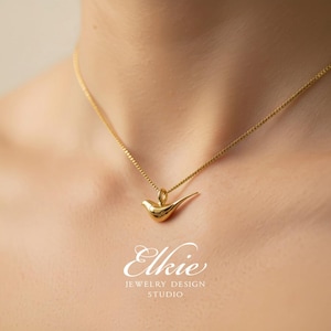 May include: A gold-toned necklace featuring a small, smooth, stylized bird pendant suspended from a delicate box chain. The pendant has a polished, reflective surface. Below the pendant, the text reads Elkie JEWELRY DESIGN STUDIO.