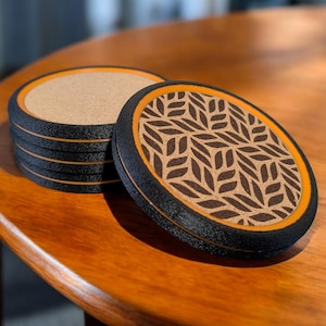 Puede incluir: Un juego de seis posavasos redondos. Cada posavasos tiene un borde negro, un anillo interior naranja y un centro de corcho. El posavasos superior presenta un patrón de hojas marrones. Los posavasos están apilados sobre una mesa de madera.