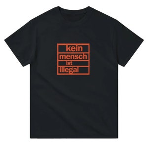 Könnte beinhalten: Schwarzes T-Shirt mit einer orangenen Grafik, auf der in einem Kasten "kein mensch ist illegal" steht.