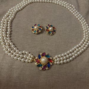 Könnte beinhalten: Ein dreireihiges Perlenketten- und Ohrring-Set. Die Halskette hat einen goldenen Verschluss mit einem zentralen Blumendesign, das mit farbigen Edelsteinen und einer großen Perle verziert ist. Die Ohrringe haben ein ähnliches Blumendesign.