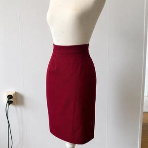 May include: A solid deep red high-waisted pencil skirt displayed on a mannequin. The skirt features a clean, structured silhouette with a fitted waistband and a straight hemline that falls just below the knee, showcasing a smooth, uniform fabric texture.