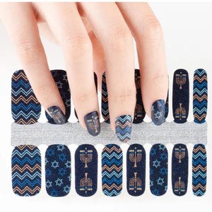 May include: Nail wraps with a navy blue background and various designs. Some wraps feature a gold menorah, others a blue and orange chevron pattern, and some have a six-pointed star. The wraps are shown with a hand.