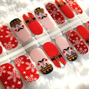 May include: Set of 10 nail wraps featuring a red and white snowflake design with a glitter finish and a nutcracker design with a red and gold uniform.