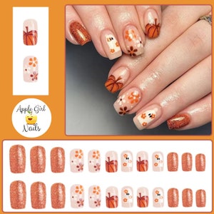 May include: Set of 10 press-on nails with a fall theme. The nails feature a variety of designs including pumpkins, flowers, and glitter. The nails are in various shades of orange, brown, and white.
