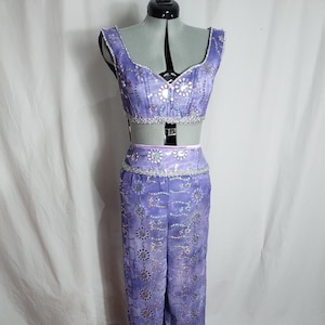 Puede incluir: Un conjunto de dos piezas color lavanda que consta de un top corto sin mangas y pantalones de pierna ancha y cintura alta. La tela presenta un patrón circular repetitivo con detalles metálicos plateados y lentejuelas en todo el conjunto.