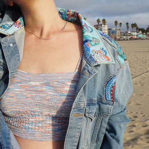 May include: A blue denim jacket with a colorful patterned collar and embroidered details. The jacket is worn over a striped knit crop top.