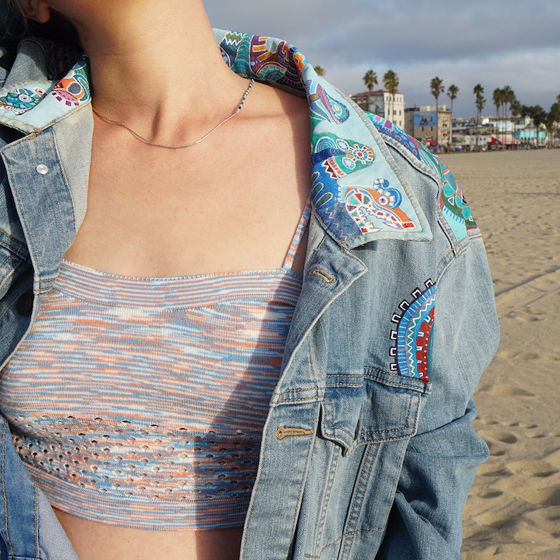 May include: A blue denim jacket with a colorful patterned collar and embroidered details. The jacket is worn over a striped knit crop top.