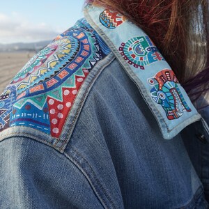 May include: A denim jacket with colorful, geometric patterns painted on the collar and shoulder. The patterns are a mix of blue, green, red, yellow, and white.