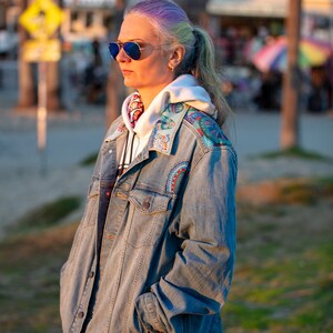 May include: A woman with purple and gray hair is wearing a blue denim jacket with colorful embroidery on the collar and shoulders. She is also wearing a white hoodie and sunglasses.