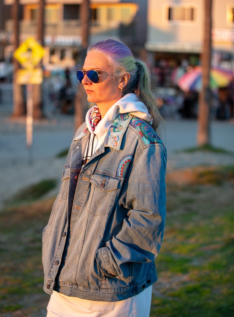 May include: A woman with purple and gray hair is wearing a blue denim jacket with colorful embroidery on the collar and shoulders. She is also wearing a white hoodie and sunglasses.