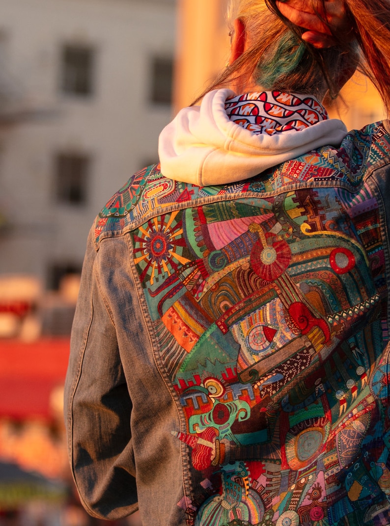 May include: A denim jacket with a colorful, abstract design painted on the back. The design features geometric shapes, swirls, and lines in shades of red, blue, green, yellow, and orange.