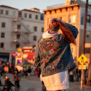 May include: A person wearing a denim jacket with a colorful, abstract design on the back. The jacket is worn over a white hoodie and a white skirt. The person is standing in front of a building with a staircase.
