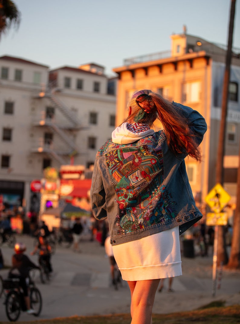 May include: A person wearing a denim jacket with a colorful, abstract design on the back. The jacket is worn over a white hoodie and a white skirt. The person is standing in front of a building with a staircase.