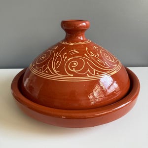 May include: A terracotta tagine pot with a conical lid and a circular base. The lid features a cream-colored, ornate, swirling design. The pot is a rich, reddish-brown color and sits on a white surface.