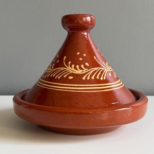 May include: A terracotta tagine pot, featuring a conical lid and a broad, shallow base. The lid is adorned with cream-coloured floral patterns and horizontal bands. The tagine is reddish-brown and rests on a white surface.