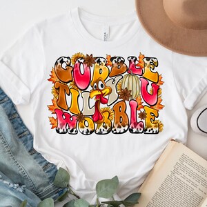 May include: White t-shirt with a Thanksgiving-themed graphic that says "Gobble Til You Wobble" with a cartoon turkey, fall leaves, and sunflowers.
