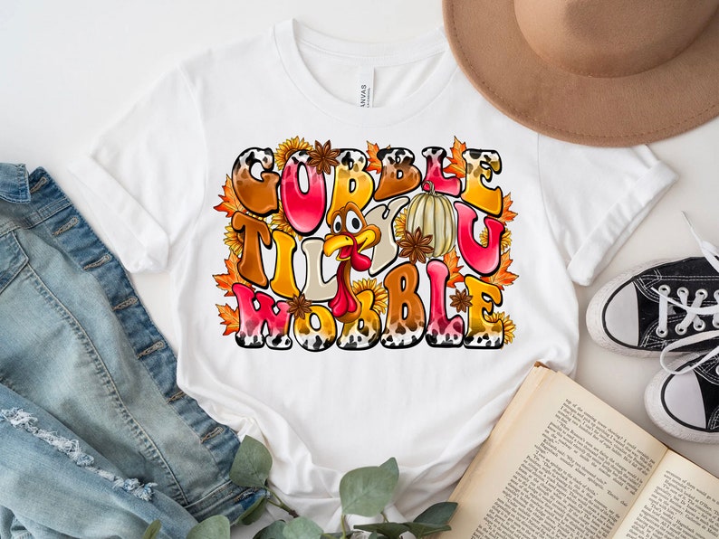 May include: White t-shirt with a Thanksgiving-themed graphic that says "Gobble Til You Wobble" with a cartoon turkey, fall leaves, and sunflowers.