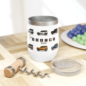 Puede incluir: Un vaso de acero inoxidable blanco con una tapa plateada y un diseño de diferentes vehículos Ford Bronco de colores. El texto "The Bronco Shop" está impreso en el vaso.