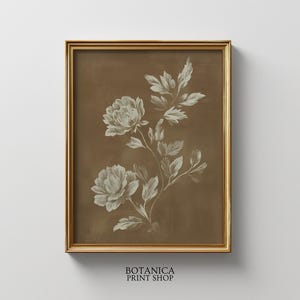 以下が含まれることがあります： 茶色の背景に白い花と葉の詳細なイラストが描かれた、額入りの植物プリント。アートワークは金のフレームに入っており、下部に「BOTANICA PRINT SHOP」というテキストが含まれています。