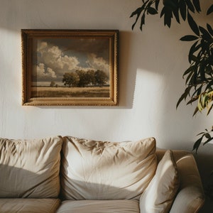 May include: A beige couch with two pillows and a throw pillow. A framed landscape painting hangs on the wall behind the couch. A green plant is in the corner of the room.