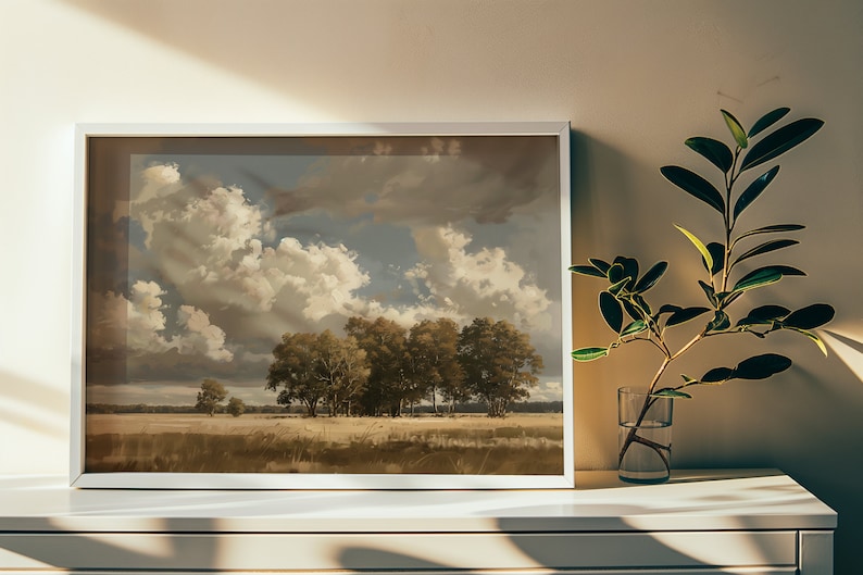 May include: A framed landscape painting of a field with trees and a cloudy sky. The painting is in a white frame and is hanging on a white wall.