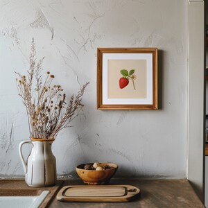 May include: A framed botanical illustration of a single red strawberry with green leaves and white flowers on a beige background. The frame is made of wood and has a light brown finish.