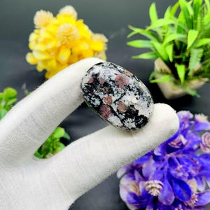 May include: A smooth, oval-shaped stone with a white, black, and pink speckled pattern. The stone is being held in a hand.