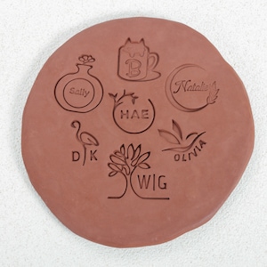 Puede incluir: Un primer plano de una losa de arcilla terracota con varios dise&ntilde;os tallados en ella. Los dise&ntilde;os incluyen una taza con un gato dentro, un jarr&oacute;n con una flor, una luna creciente con el nombre "Natalie", un &aacute;rbol con el nombre "WIG", un flamenco con el nombre "DJ", una hoja con el nombre "OLIVIA", y un c&iacute;rculo con el nombre "HAE".