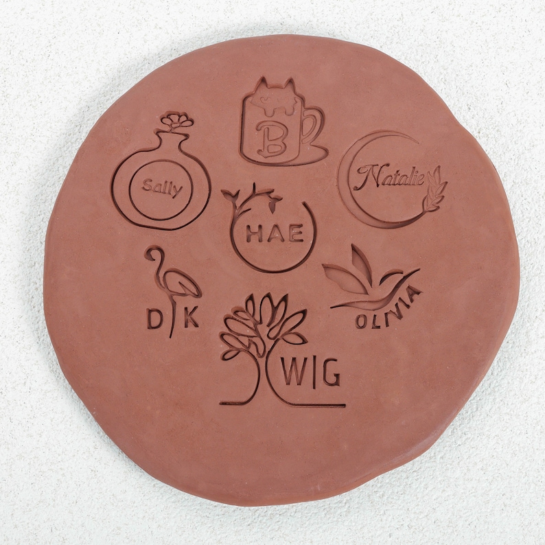 Puede incluir: Un primer plano de una losa de arcilla terracota con varios dise&ntilde;os tallados en ella. Los dise&ntilde;os incluyen una taza con un gato dentro, un jarr&oacute;n con una flor, una luna creciente con el nombre "Natalie", un &aacute;rbol con el nombre "WIG", un flamenco con el nombre "DJ", una hoja con el nombre "OLIVIA", y un c&iacute;rculo con el nombre "HAE".