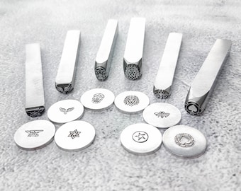 May include: A set of 12 metal stamps with various designs, including a skull, a lion, a bee, a star, and a spade. The stamps are arranged in two rows on a gray surface.