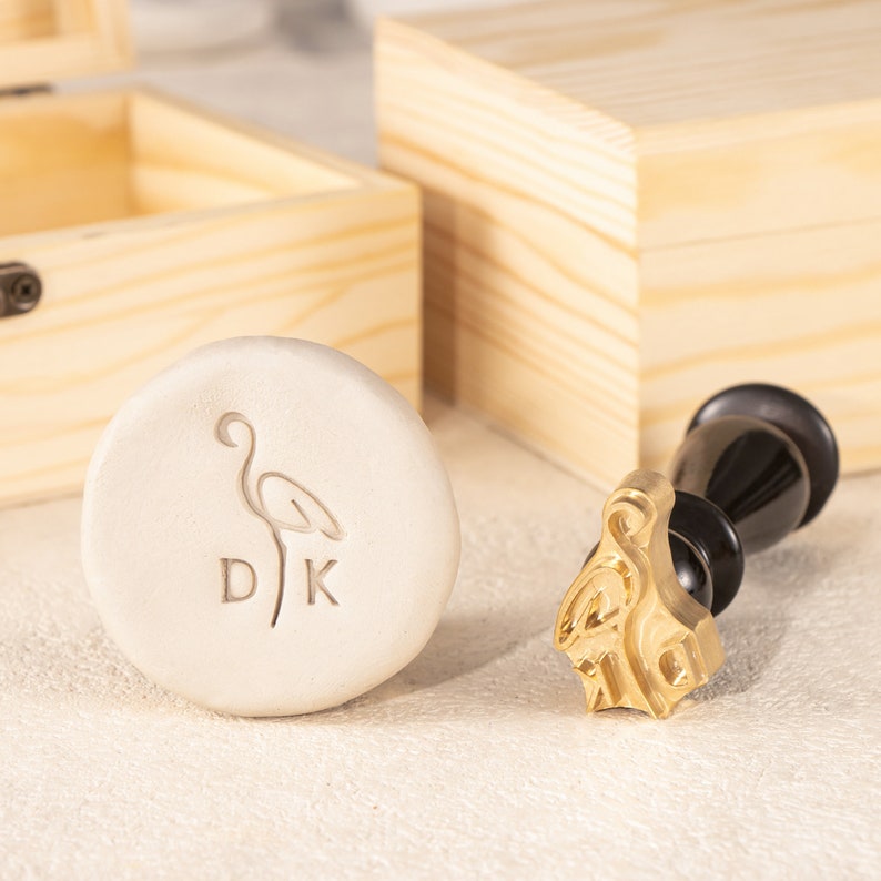 Puede incluir: Un sello de metal dorado con un dise&ntilde;o de flamenco y las letras "DK" sobre una superficie de arcilla blanca. El sello est&aacute; unido a un mango de madera negro.