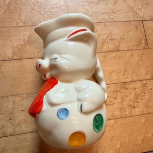 May include: A cream-colored ceramic pitcher shaped like a pig wearing a sailor hat. The pig has painted black eyelashes, a red scarf, and colorful blue, green, and orange polka dots on its round body. It features a curved handle on the side.