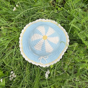 Puede incluir: Un sombrero de sol de crochet con un diseño de margarita en tonos de azul, blanco y amarillo. El sombrero está sobre un lecho de hierba verde.
