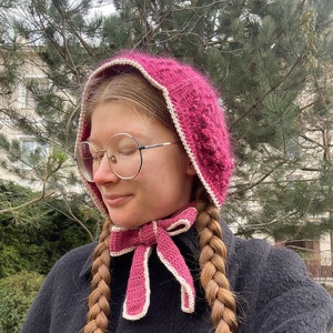 May include: A woman wearing a pink crocheted hat with a white trim and a matching pink and white crocheted scarf. She is wearing round glasses and has her hair in two braids.