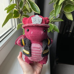 May include: A crocheted pink dragon wearing a firefighter's hat and jacket. The dragon has black eyes and a grey helmet.