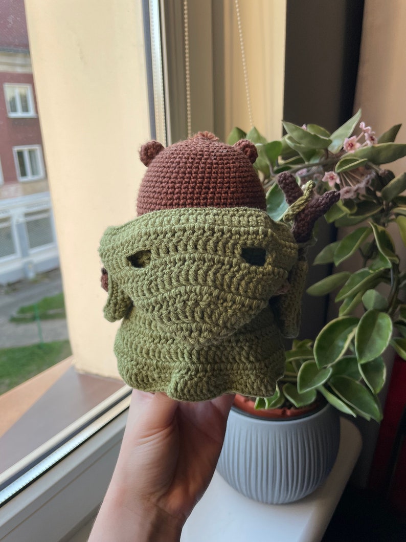 May include: A crocheted stuffed toy with a brown head and green body. The toy is wearing a green crocheted cloak with a brown crocheted hat. The toy is being held in a person's hand.